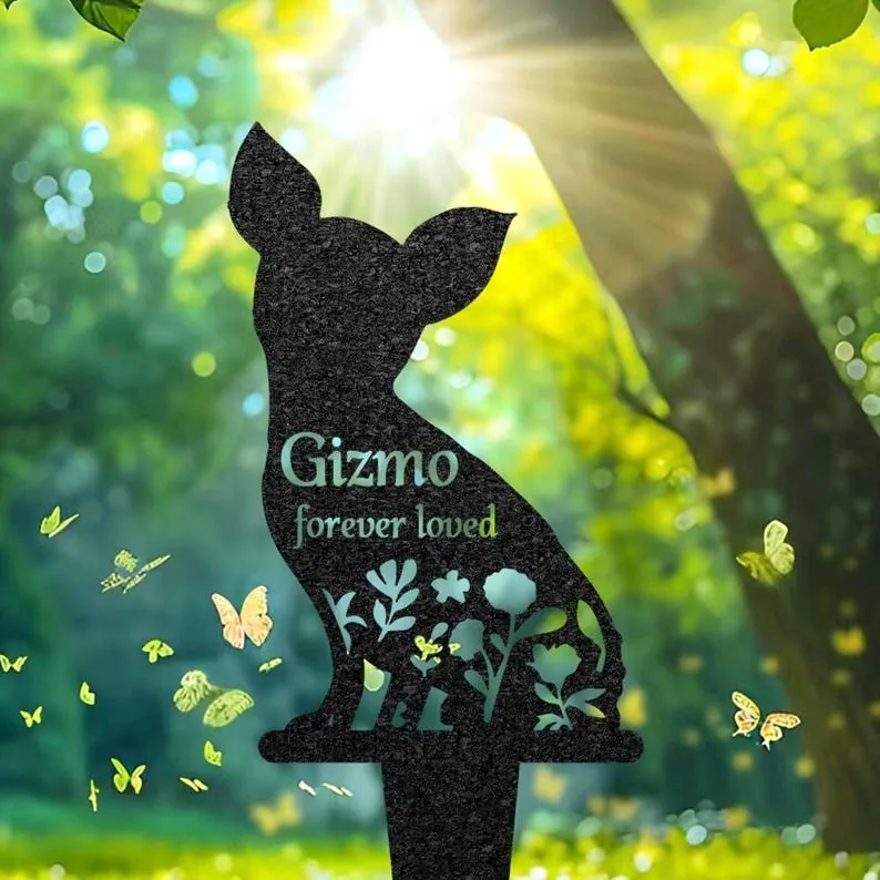 Silhouette garden memorial stake shaped like a small dog with floral cutouts and text 'Gizmo forever loved' in a sunlit wooded yard