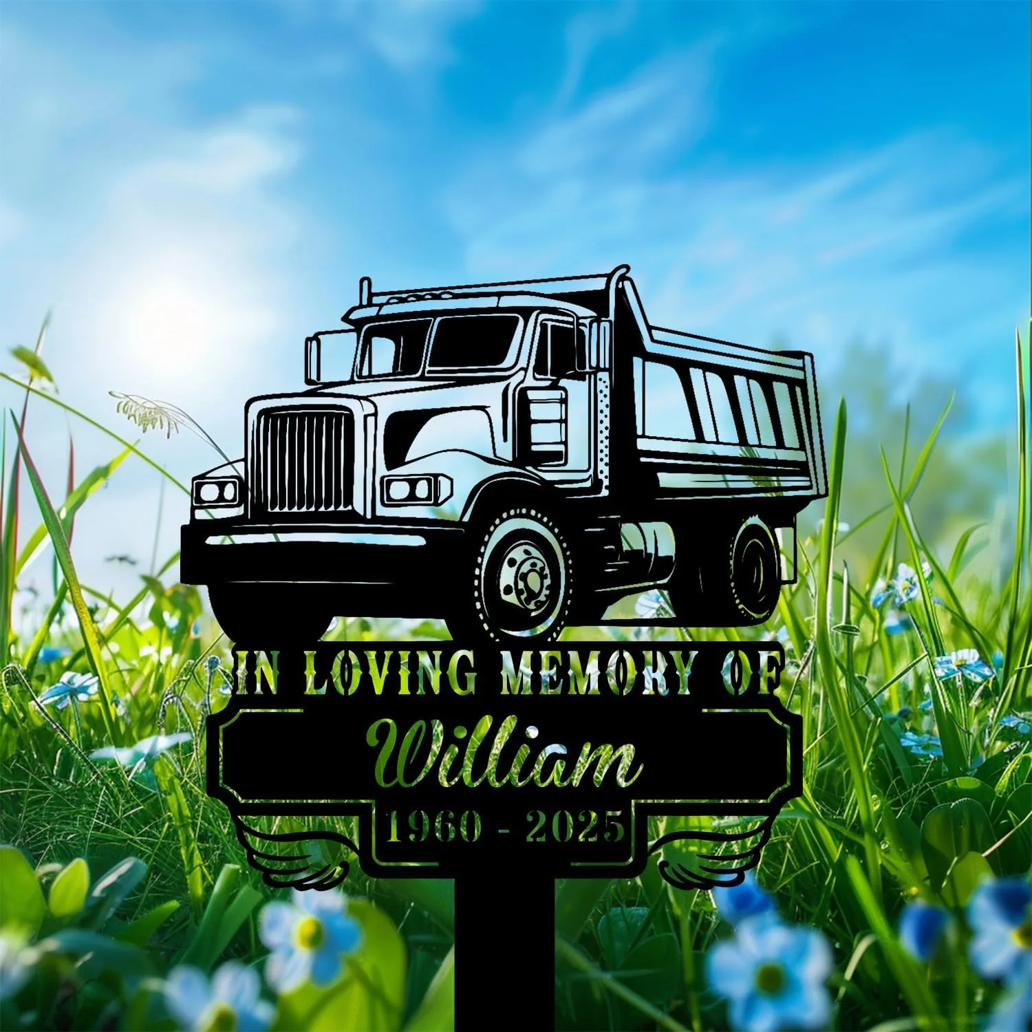 Black silhouette memorial sign showing a dump truck above text "In Loving Memory of William 1960 - 2025" placed among green grass and small blue flowers under a bright blue sky