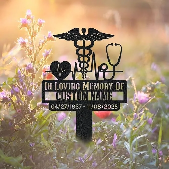 Black memorial garden stake with caduceus, stethoscope, heartbeat and heart icons reading "In Loving Memory of CUSTOMNAME" and dates 04/27/1967 - 11/08/2025 among wildflowers