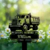 Black silhouette memorial sign shaped like a dump truck reading "In Loving Memory of William 1960 - 2025" placed among daisies in a sunlit green field
