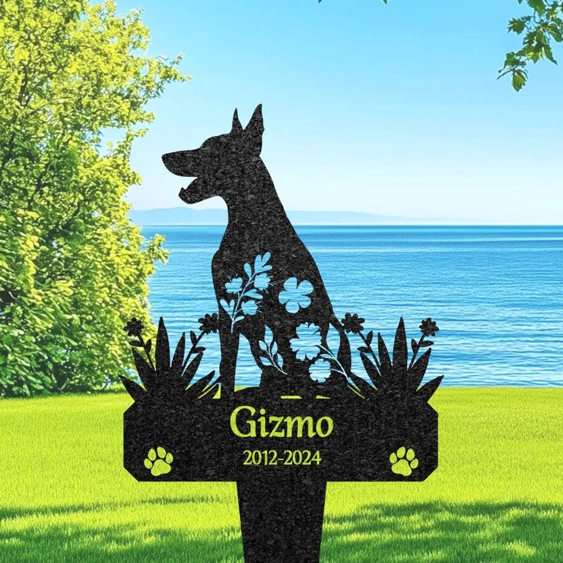 Black metal dog silhouette grave marker reading “Gizmo 2012-2024” with floral cutouts and paw prints set on green lawn by a lakeside