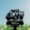 Black memorial sign with a dump truck illustration and text "In Loving Memory of William 1960 - 2025" set against a grassy field of dandelions and a blue sky