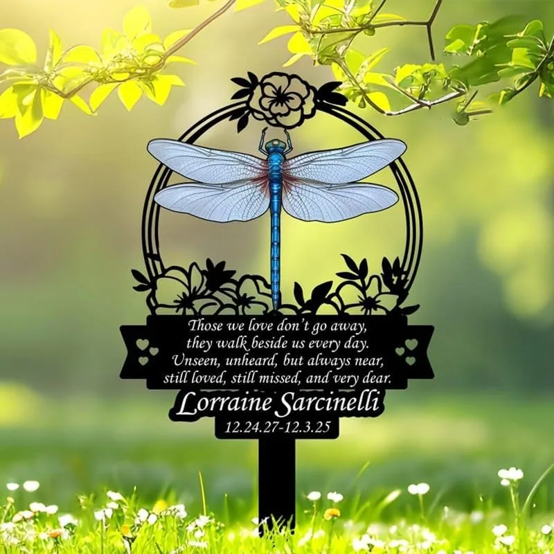 Garden memorial stake with a blue dragonfly and floral circle, engraved poem and name 'Lorraine Sarcinelli' with dates 12.24.27–12.3.25, placed in grassy yard