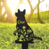 Black metal garden stake in the silhouette of a sitting dog with floral cutouts and the name “Coco” in gold, placed in a sunlit grassy yard