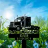 Black silhouette memorial sign of a semi truck with text "In Loving Memory Of" and "DAD 1949-2024" set among green grass and blue sky with small blue flowers