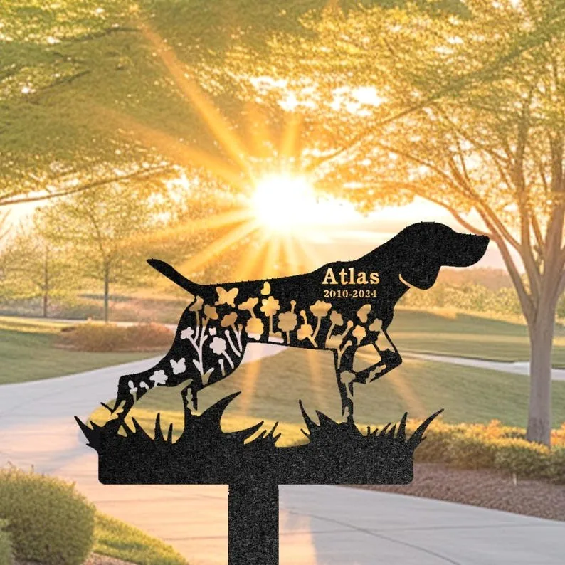 Black metal dog silhouette memorial yard stake with floral cutouts and engraved name 'Atlas 2010-2024', placed outdoors in a sunlit park-like yard