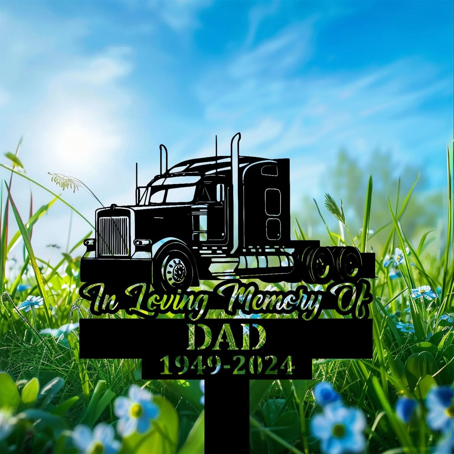 Black silhouette memorial sign of a semi truck with text "In Loving Memory Of" and "DAD 1949-2024" set among green grass and blue sky with small blue flowers