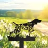 Black metal dog silhouette garden stake with floral cutouts and engraved name "Atlas 2010-2024" against a sunny meadow background