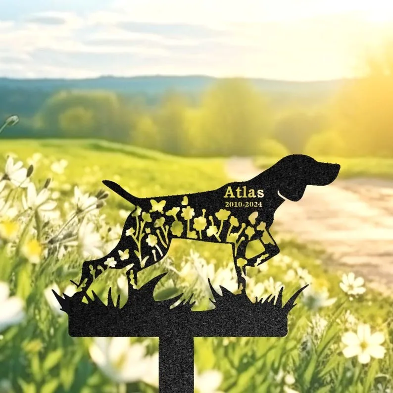 Black metal dog silhouette garden stake with floral cutouts and engraved name "Atlas 2010-2024" against a sunny meadow background