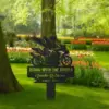 Black memorial garden stake showing a winged motorcyclist silhouette and text "Riding With The Angels, Amalin Robinson, 1962 - 2025" placed in a grassy park with tulip beds and trees