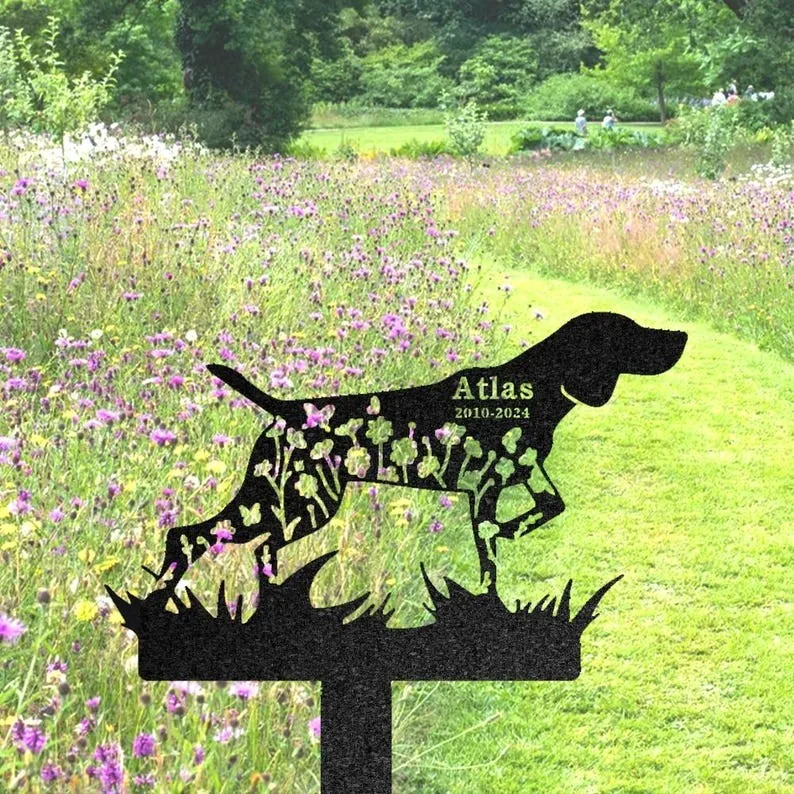Black metal dog silhouette garden stake with floral cutouts and engraved name "Atlas" and years "2010-2024" placed in a meadow of purple wildflowers