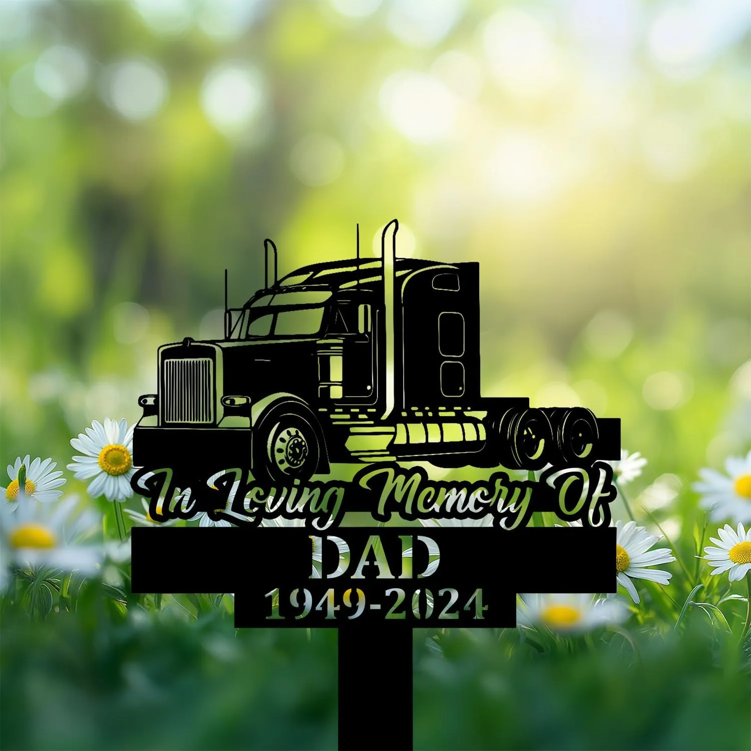 Black memorial yard sign with a semi truck silhouette reading "In Loving Memory Of" and "DAD 1949-2024" placed among daisies in a sunlit garden