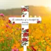 White memorial cross with red rose illustrations reading "In celebration of a beautiful life Emily 1963-2024" placed in a sunlit wildflower field