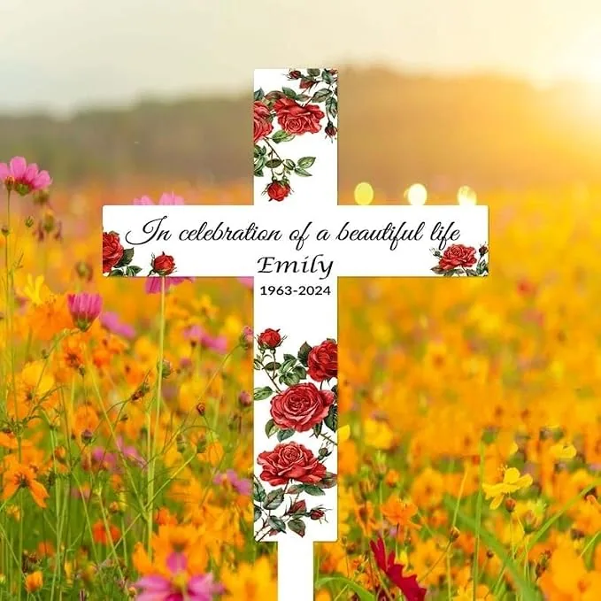 White memorial cross with red rose illustrations reading "In celebration of a beautiful life Emily 1963-2024" placed in a sunlit wildflower field