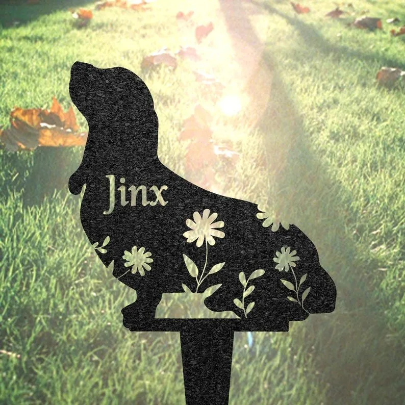 Black silhouette garden stake shaped like a dog with cutout flowers and the name "Jinx", placed in sunlit grassy yard