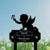 Black silhouette memorial sign of a winged baby reaching for a butterfly above text 'I'll hold you in my heart until I can hold you in heaven 2026' set against a blue sky and dandelion field
