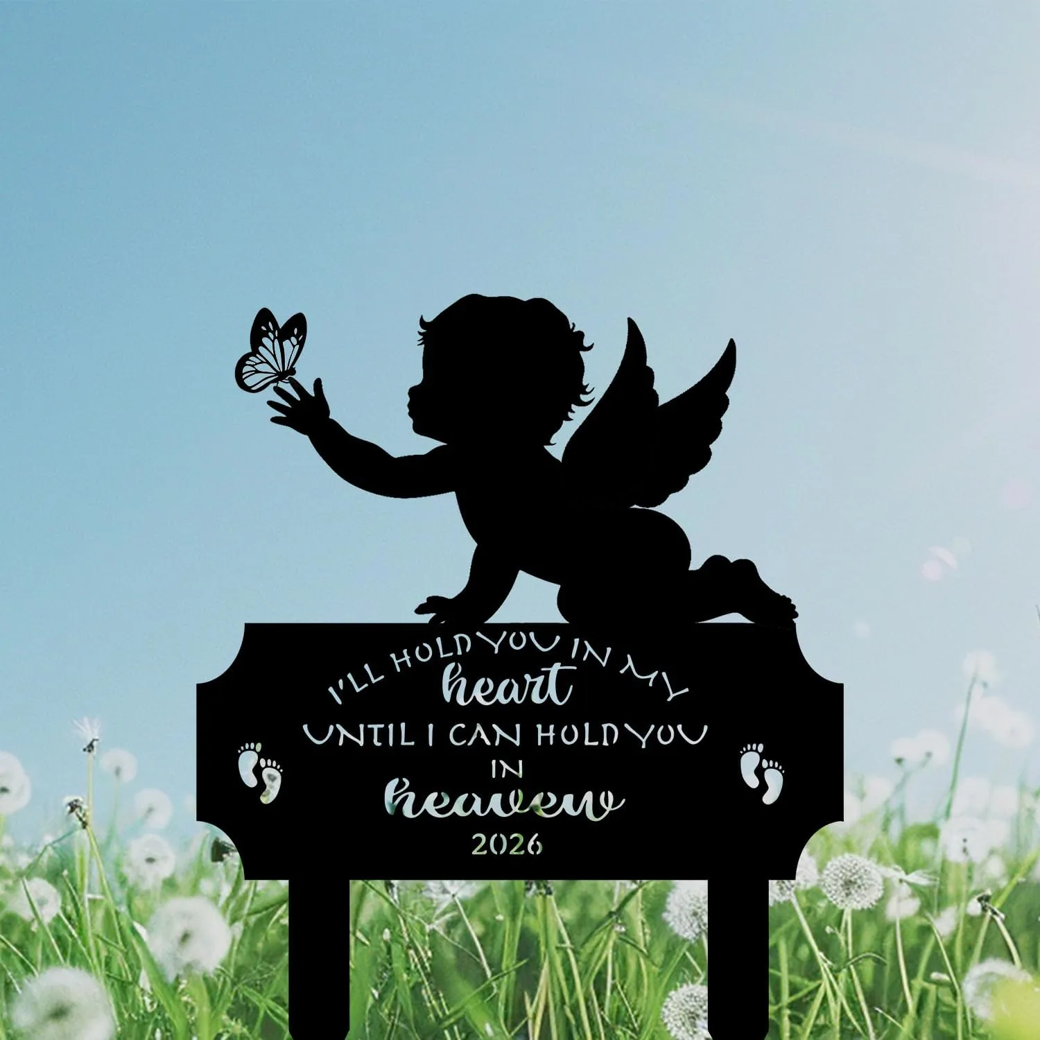 Black silhouette memorial sign of a winged baby reaching for a butterfly above text 'I'll hold you in my heart until I can hold you in heaven 2026' set against a blue sky and dandelion field