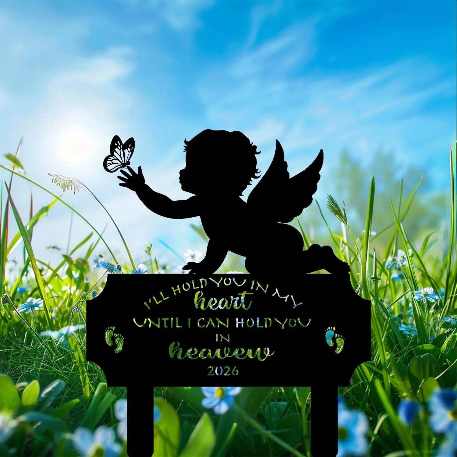 Silhouette of a winged baby reaching for a butterfly above a memorial plaque in a sunny grassy field with blue sky and wildflowers, text reads "I'll hold you in my heart until I can hold you in heaven 2026"