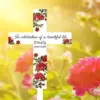 White memorial cross with red rose pattern reading "In celebration of a beautiful life Emily 1963-2024" set against a sunlit garden background with pink roses