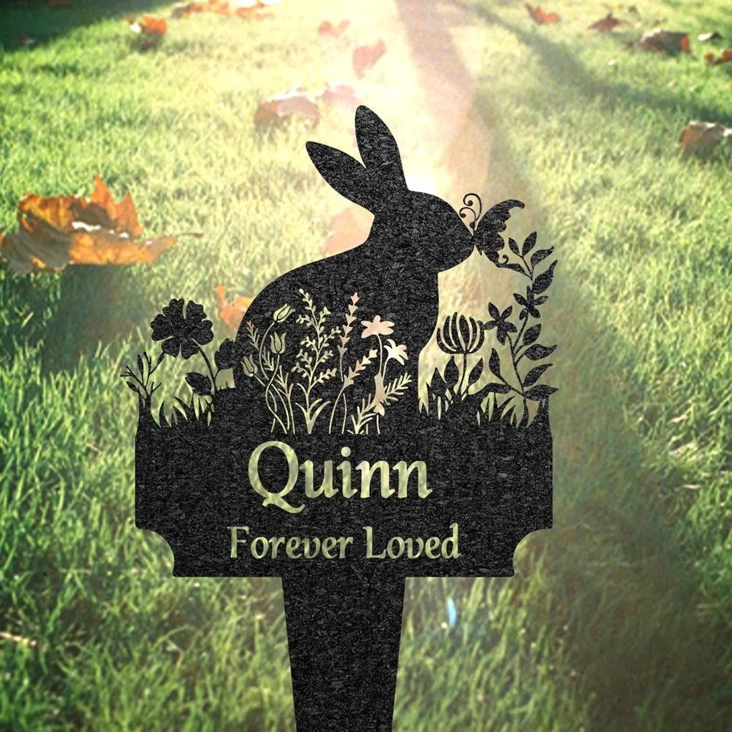 32-2 Black metal garden memorial stake shaped like a rabbit with floral cutouts and text reading "Quinn Forever Loved" placed in sunlit grass