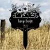 Black memorial garden stake in a grassy field reading “Planted In Loving Memory Of Lucy Swift 1980–2021 Isaiah 12:2” with floral illustration at top
