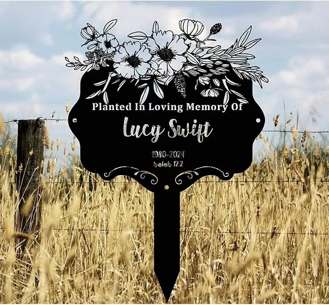 Black memorial garden stake in a grassy field reading “Planted In Loving Memory Of Lucy Swift 1980–2021 Isaiah 12:2” with floral illustration at top