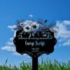Black memorial garden sign reading “Planted In Loving Memory Of Lucy Swift 1980–2024 Isaiah 12:2” with floral illustration against a blue sky and grass