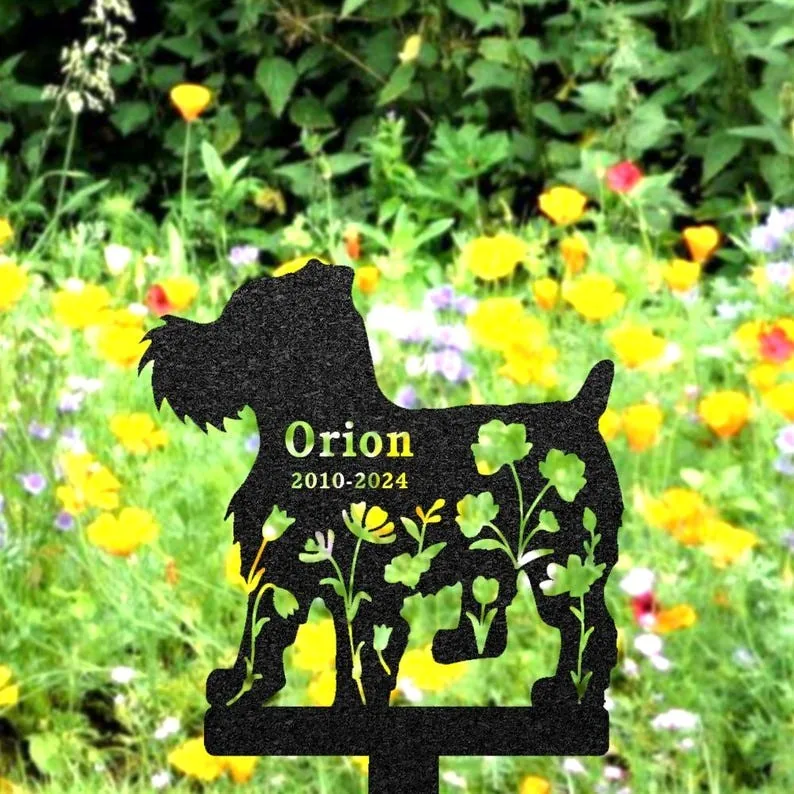 Black metal garden memorial silhouette of a small dog with cutout flowers and text "Orion 2010-2024" staked in a colorful wildflower meadow