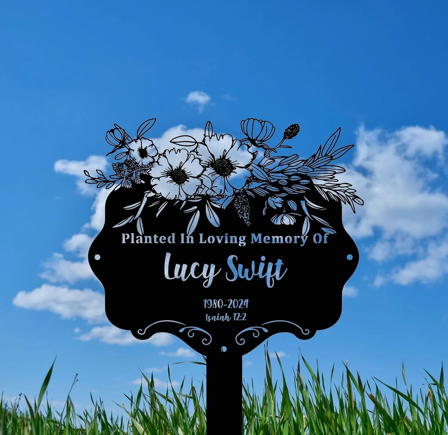 Black memorial garden sign reading “Planted In Loving Memory Of Lucy Swift 1980–2024 Isaiah 12:2” with floral illustration against a blue sky and grass