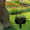 Black memorial garden sign reading "Planted In Loving Memory Of Lucy Swift" staked in grassy park bed with tulips and a tree trunk nearby