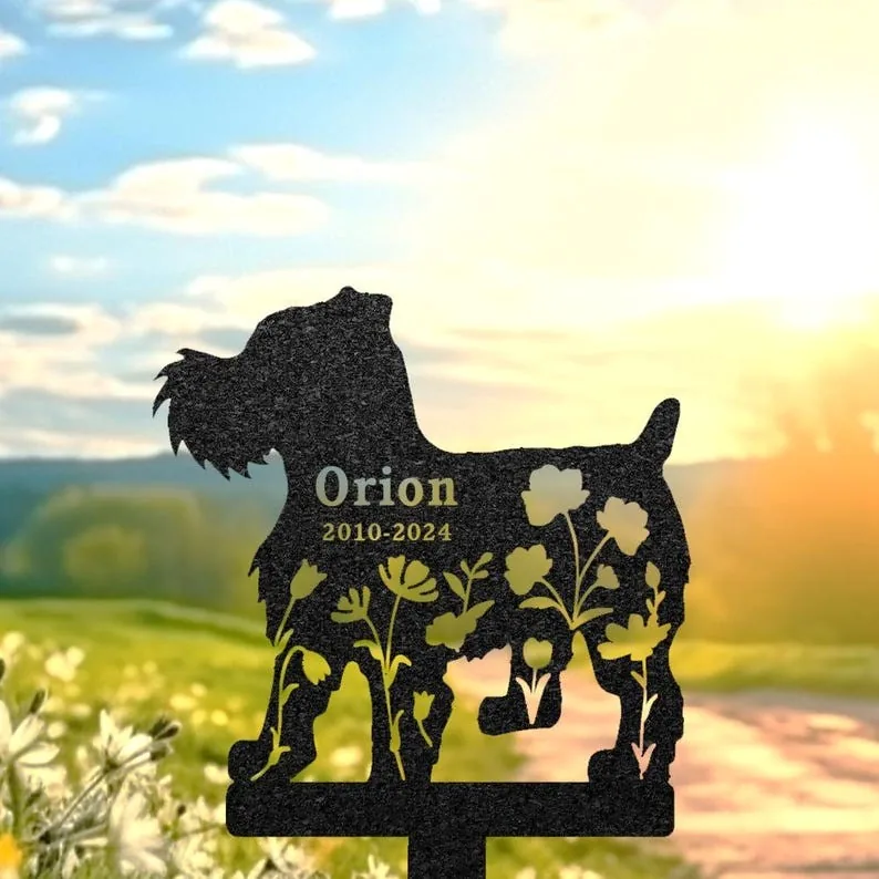 Black metal dog-shaped memorial stake with floral cutouts and text "Orion 2010-2024" placed outdoors in a sunny meadow at sunset