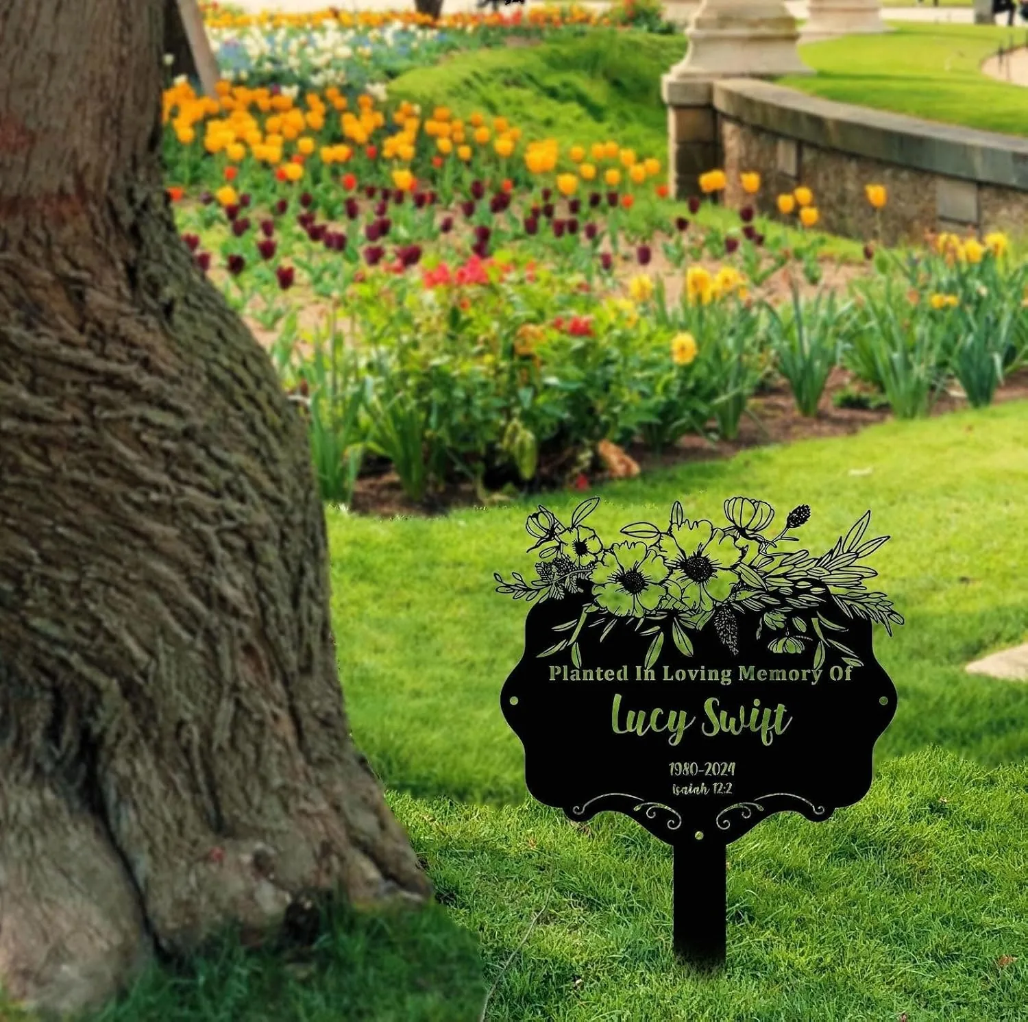 Black memorial garden sign reading "Planted In Loving Memory Of Lucy Swift" staked in grassy park bed with tulips and a tree trunk nearby