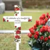 White memorial cross sign with red rose decorations reading “In celebration of a beautiful life Emily 1963-2024” placed in grass beside a bouquet of red roses