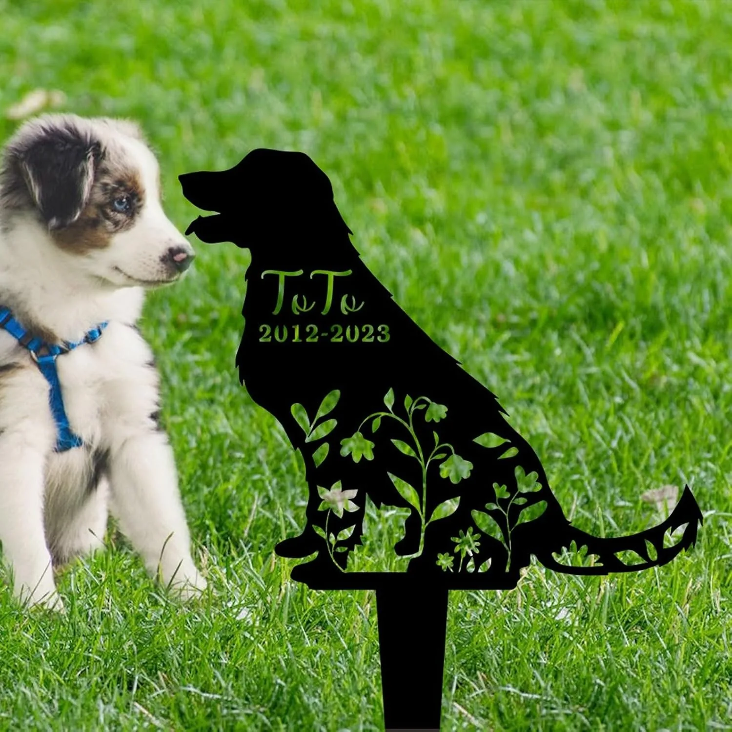Black metal dog silhouette garden memorial stake reading 'Toto 2012-2023' on grassy lawn with a puppy wearing a blue harness nearby