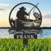 Black metal memorial yard sign showing a silhouette of a fisherman in a boat catching a fish with text "In Loving Memory" and name FRANK, set in grass by a lake at sunset