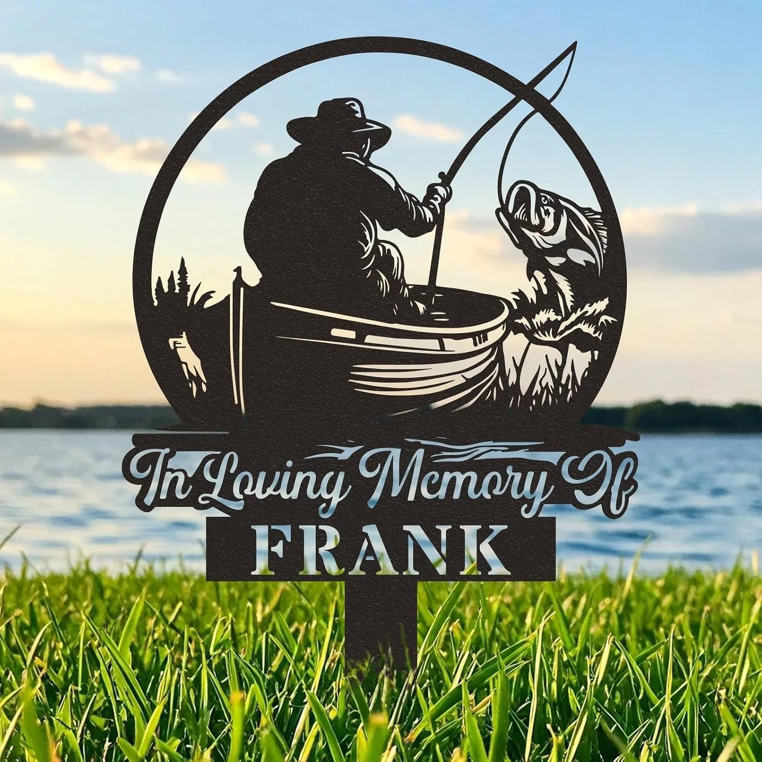 Black metal memorial yard sign showing a silhouette of a fisherman in a boat catching a fish with text "In Loving Memory" and name FRANK, set in grass by a lake at sunset