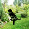 Black metal dog-shaped garden memorial stake with floral cutouts and text "TuTu 2012-2023" placed in a green backyard garden