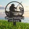 Black metal memorial yard sign reading "In Loving Memory of FRANK" with silhouette of a fisherman in a boat reeling in a fish, shown on grass by the water