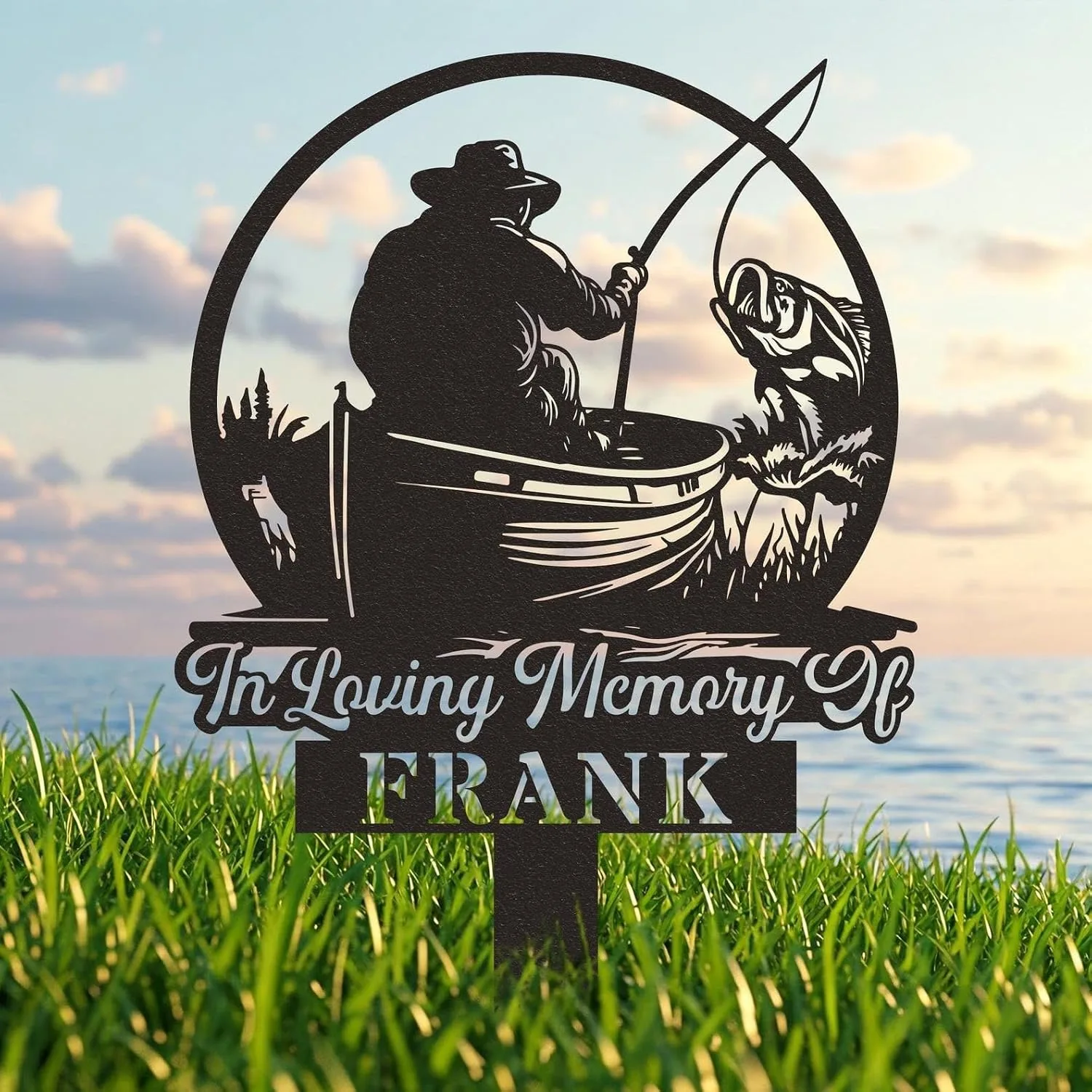 Black metal memorial yard sign reading "In Loving Memory of FRANK" with silhouette of a fisherman in a boat reeling in a fish, shown on grass by the water