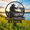 Black metal memorial stake reading "In Loving Memory FRANK" with silhouette of a fisherman in a boat catching a fish, placed in grassy lakeside at sunset