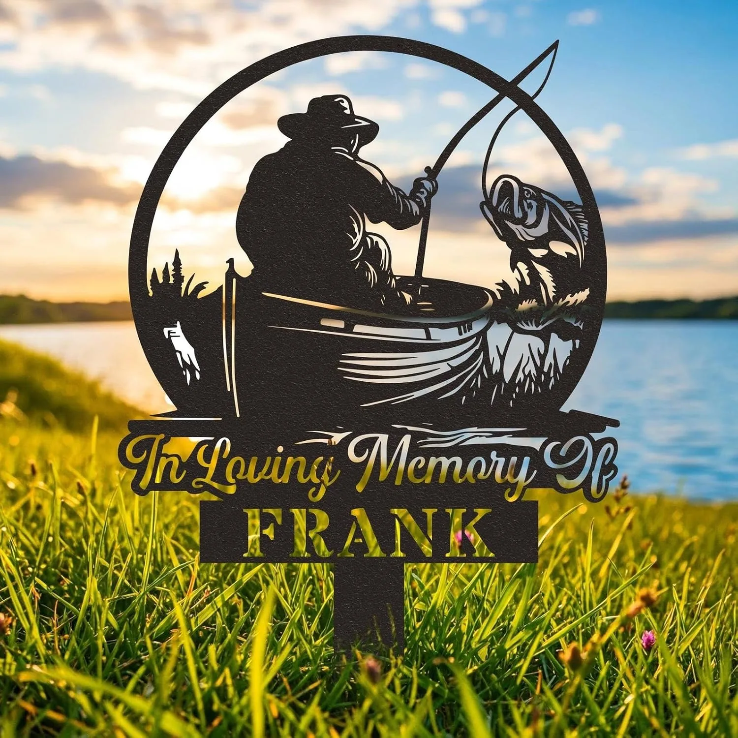 Black metal memorial stake reading "In Loving Memory FRANK" with silhouette of a fisherman in a boat catching a fish, placed in grassy lakeside at sunset