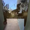 Metal silhouette pet sign reading "King Oliver" with a dachshund cutout, heart detail, and poem held in a doorway overlooking a wooden deck and outdoor string lights