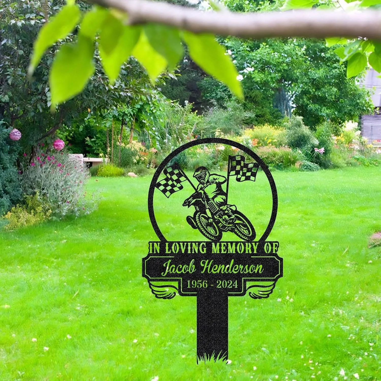 351 Black memorial lawn sign with motocross rider and checkered flags reading "In Loving Memory of Jacob Henderson 1956 - 2024" staked in a green backyard garden