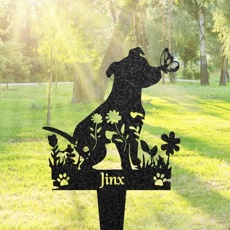 Black metal garden stake silhouette of a sitting dog with flowers, paw prints, and a butterfly, personalized with the name "Jinx", placed in a sunlit grassy park