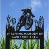Black metal memorial sign with motocross rider silhouette and text "IN LOVING MEMORY OF JACK 1987-2024" staked in grassy outdoor setting