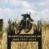 Black metal memorial yard sign showing a motocross rider silhouette and text "In Loving Memory of Jack 1987-2024" placed in a wheat field near a wire fence