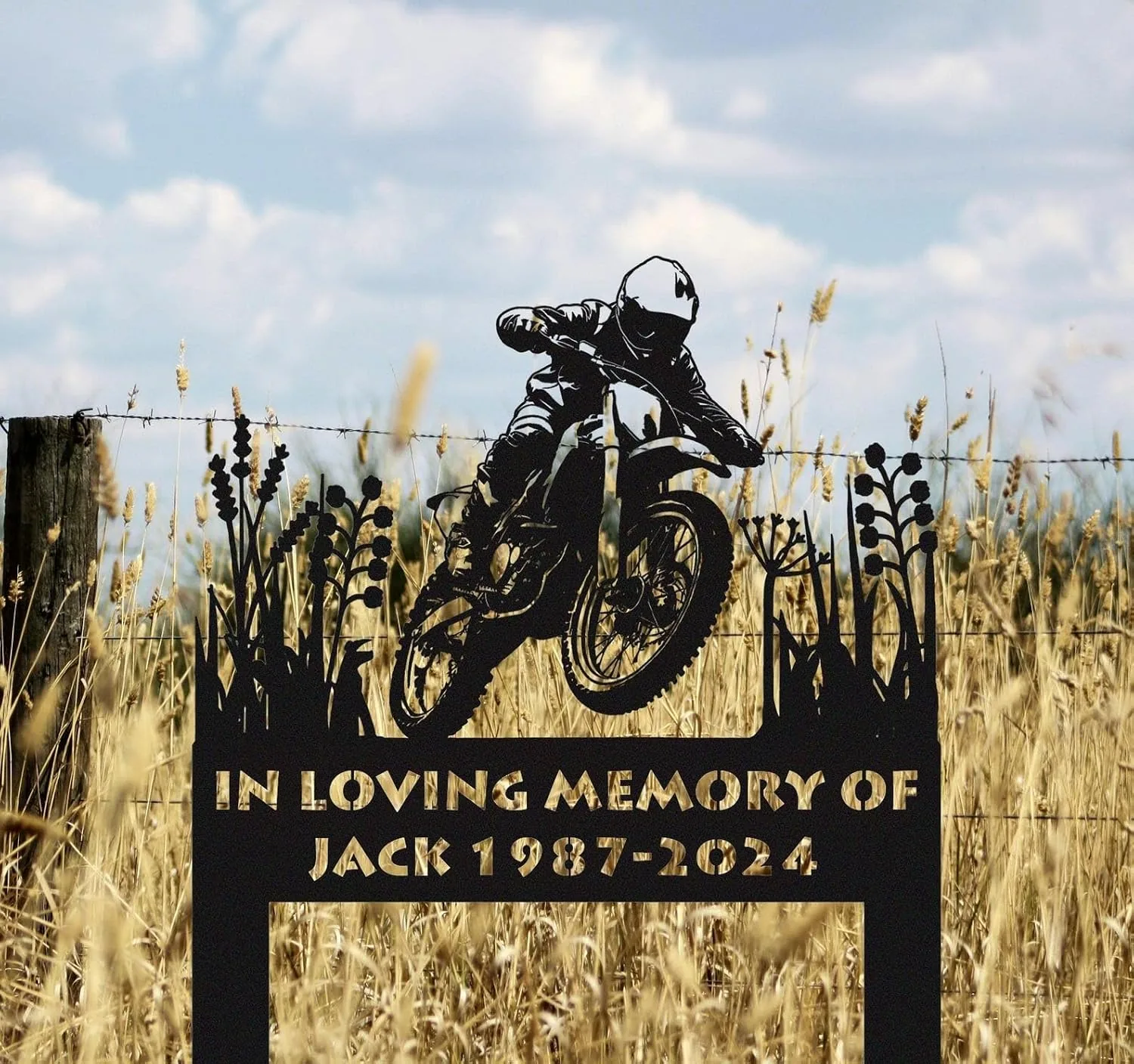 Black metal memorial yard sign showing a motocross rider silhouette and text "In Loving Memory of Jack 1987-2024" placed in a wheat field near a wire fence