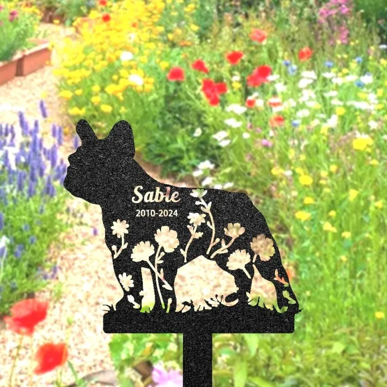 Black metal garden silhouette of a French bulldog with floral cutouts and engraved name "Sable" and dates "2010-2024" placed on a stake among wildflower garden beds