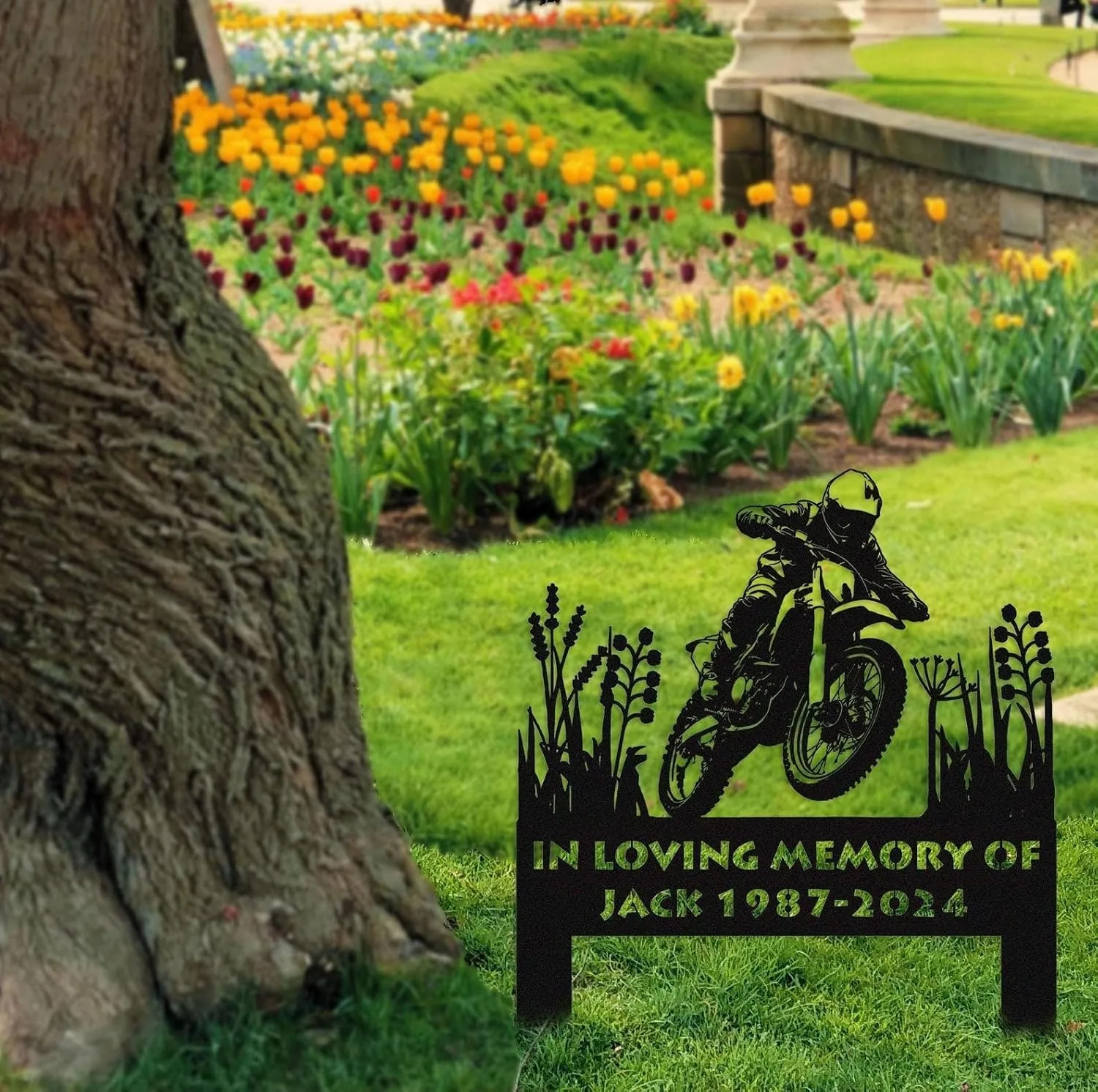 Black metal memorial lawn sign with a motocross rider silhouette and text “IN LOVING MEMORY OF JACK 1987-2024” placed on grass near a tree and colorful flowerbeds
