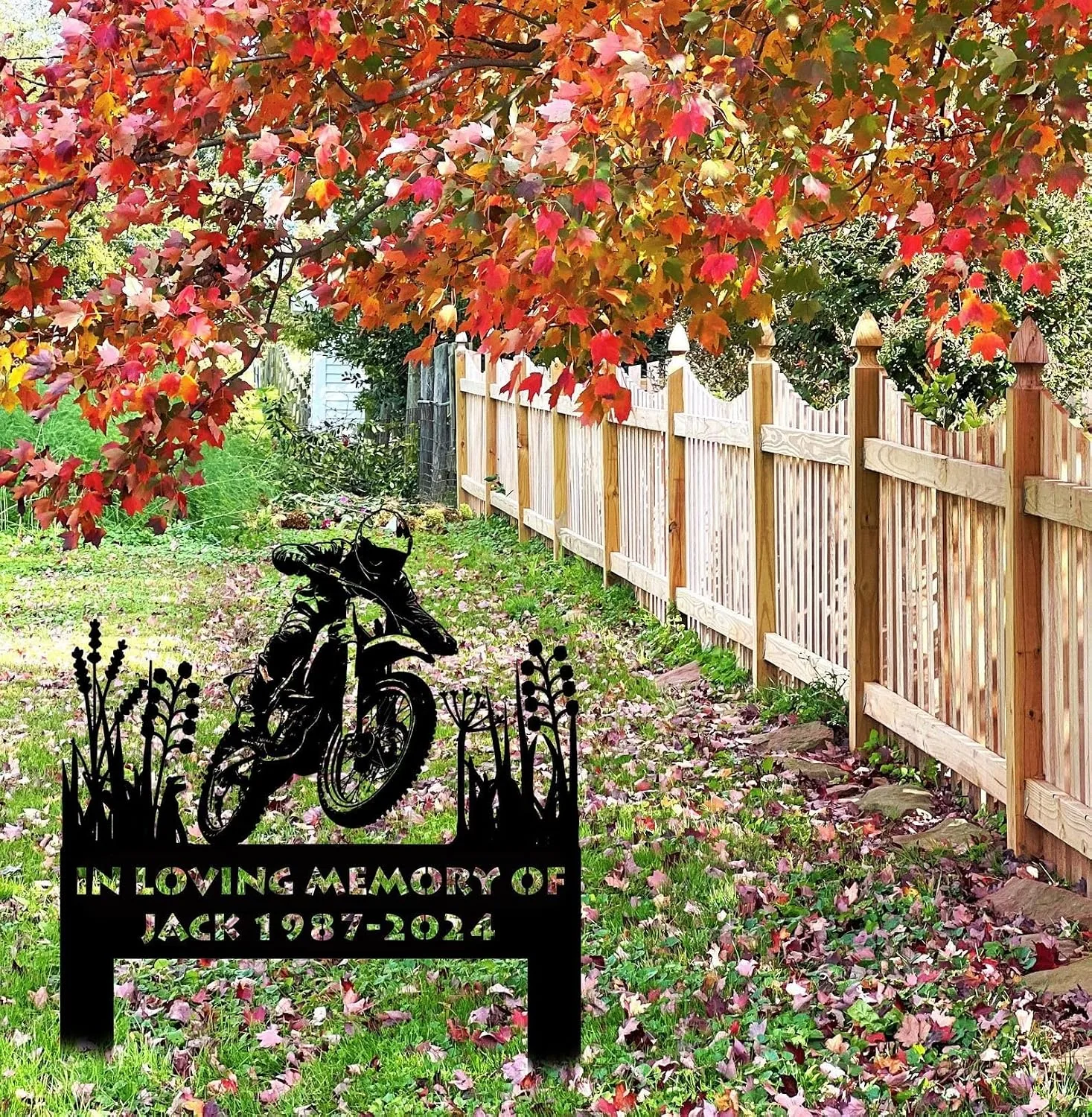 Autumn yard with red and orange tree leaves, a wooden picket fence, fallen leaves on grass, and a black metal memorial sign reading "IN LOVING MEMORY OF JACK 1987-2024" with a motorcycle silhouette.